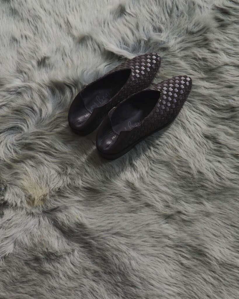 Scarpe basse Henry Beguelin Aurora Flat in pelle intrecciata scura, con punta affusolata, su una superficie grigia a pelo lungo.
