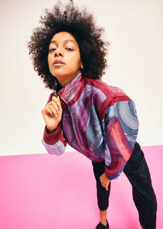 Blouson A-POC ABLE ISSEY MIYAKE x Etsu Egami bordeaux, con motivi jacquard viola e azzurri, indossato con pantaloni neri.
