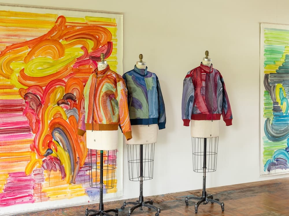 Tre giacche A-POC ABLE ISSEY MIYAKE sui manichini accanto ai dipinti di Etsu Egami, nelle varianti arancio, verde e bordeaux.