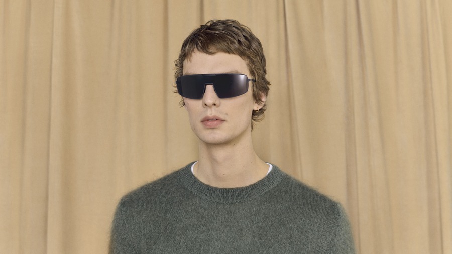 Leon Dame indossa gli occhiali a mascherina neri MYKITA RIMOWA MR008, con un maglione verde davanti a una tenda beige.