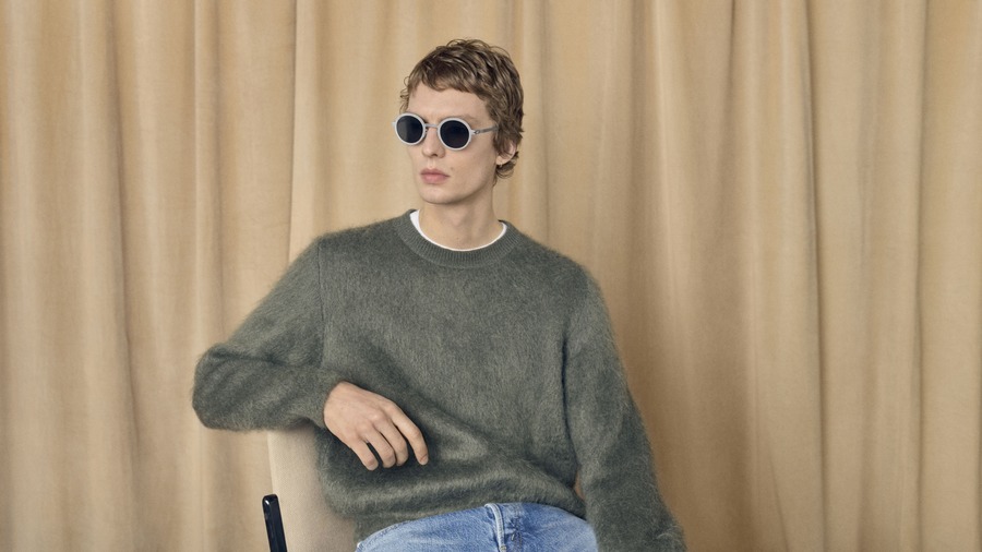 Leon Dame indossa gli occhiali rotondi MYKITA RIMOWA MR007 con montatura argento e lenti scure, abbinati a un maglione verde.