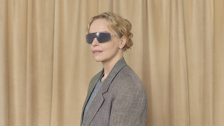 Nina Hoss indossa gli occhiali a visiera MYKITA RIMOWA MR008 con bordo argento, davanti a una tenda beige.