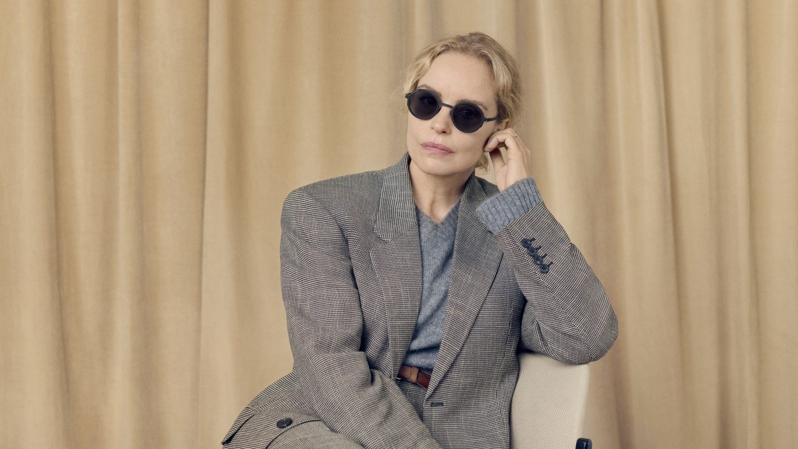 Nina Hoss indossa gli occhiali da sole rotondi neri MYKITA RIMOWA MR007, seduta davanti a una tenda beige.