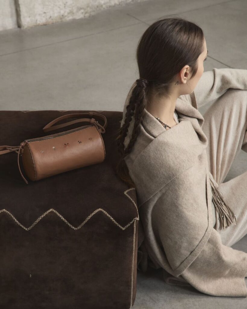 Borsa Henry Beguelin Classic Pouch Bag marrone con piccoli ricami, appoggiata su una seduta accanto a una modella in beige.