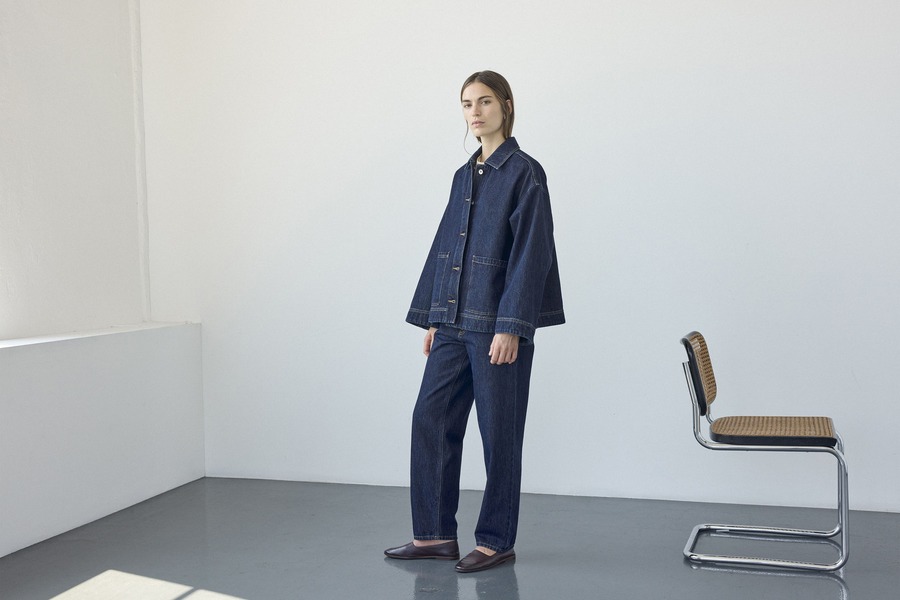 Modella con giacca ampia e pantaloni in denim indaco Pomandère, accanto a una sedia in metallo e paglia intrecciata.