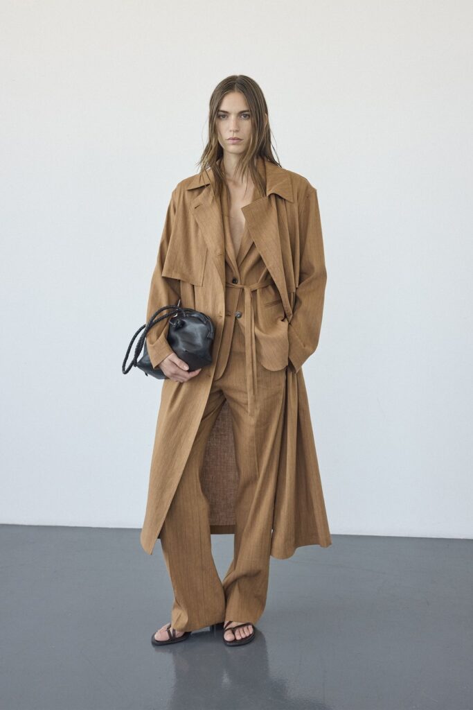 Modella con capospalla lungo, giacca e pantaloni color tabacco Pomandère, abbinati a una borsa nera e sandali.
