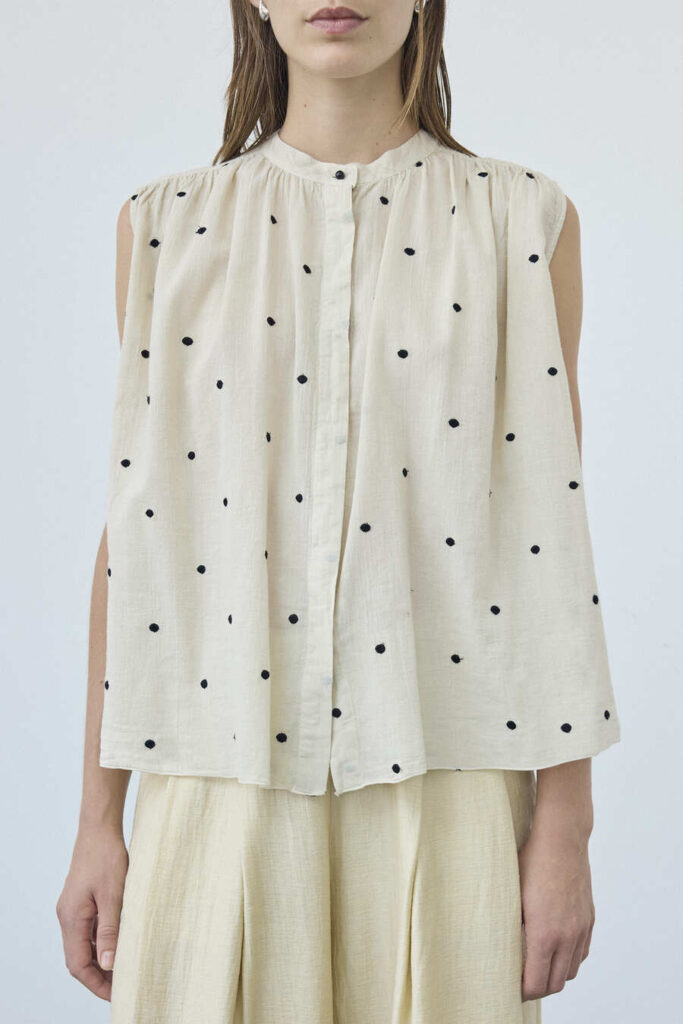 Blusa Pomandère senza maniche color avorio a piccoli pois scuri, con arricciature al collo e pantaloni chiari.