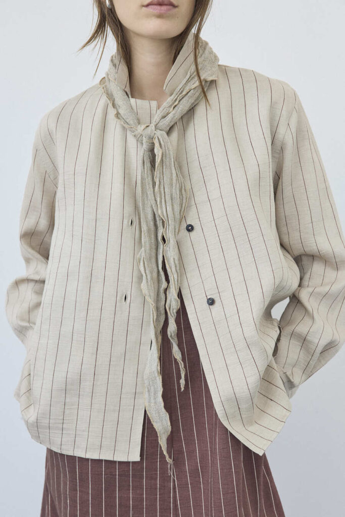 Dettaglio di un look Pomandère con giacca chiara a righe sottili, foulard annodato e capo sottostante a righe su fondo scuro.