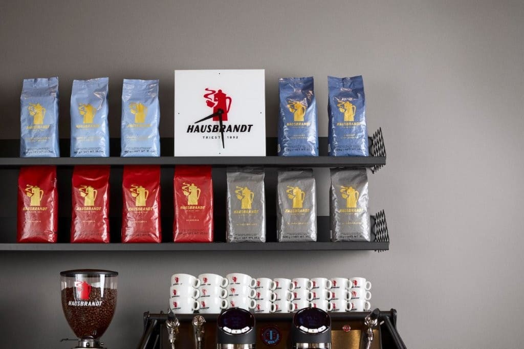 Confezioni di caffè Hausbrandt sulle mensole e tazzine sopra una macchina per espresso, con il simbolo della Moka rossa al centro.