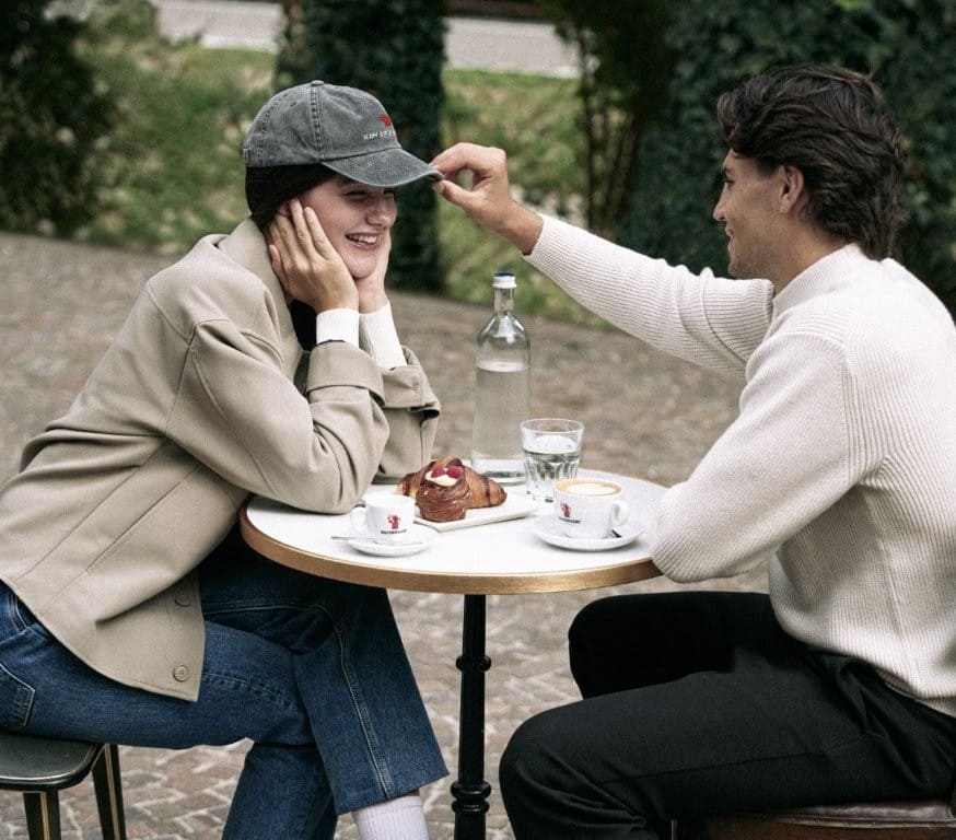 Due persone al tavolino di un bar con tazze Hausbrandt, mentre lui le sistema la visiera del cappellino.