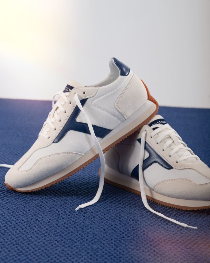 Un paio di sneakers Testoni bianche con inserti blu, lacci bianchi e suola color gomma, appoggiate su una superficie blu.