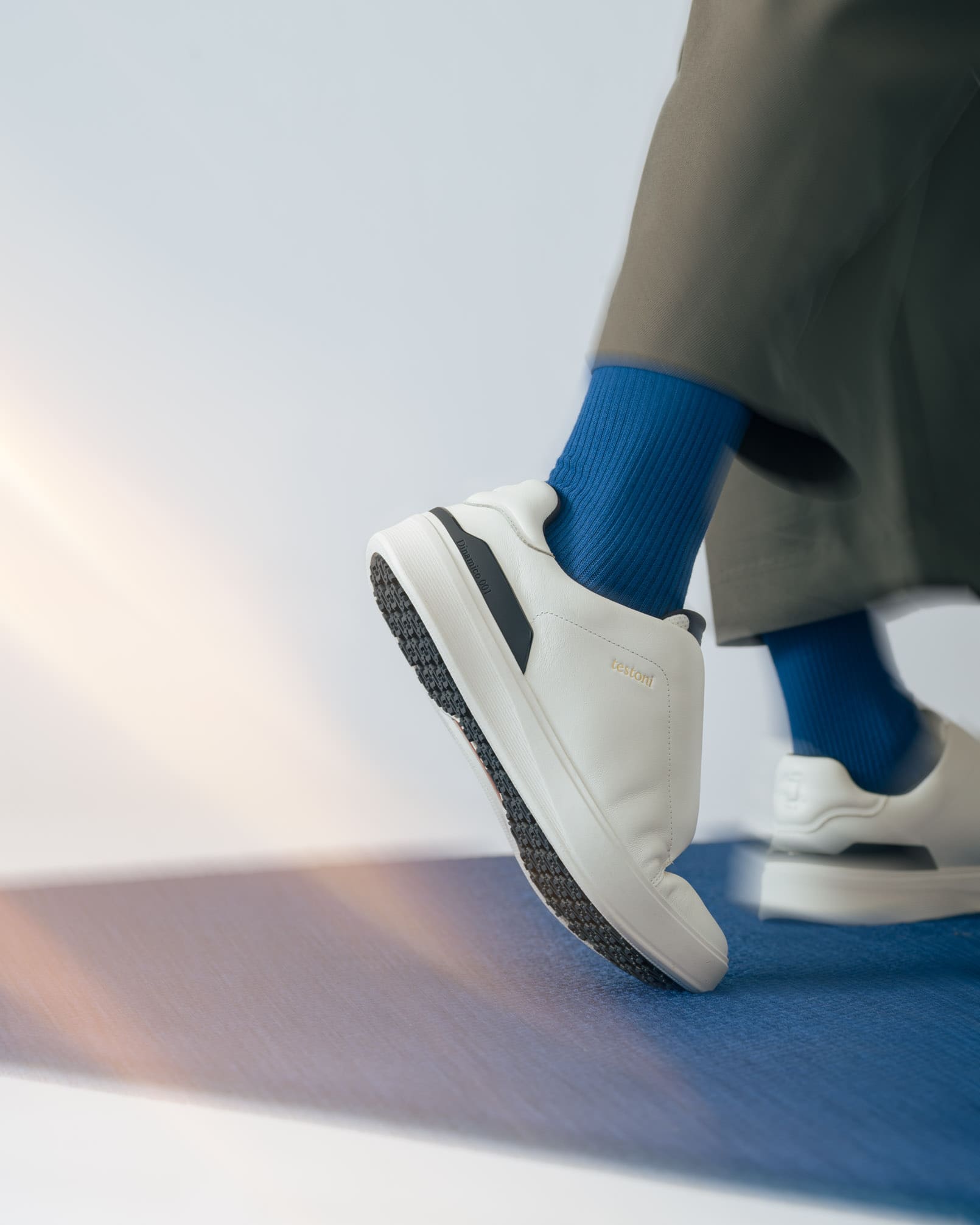 Sneakers Testoni Dinamico 001 bianche senza lacci, con dettaglio scuro sul tallone, indossate con calze blu e pantaloni grigi.